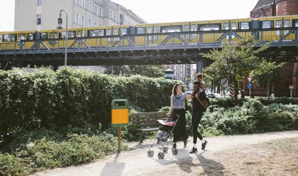 Young man carrying daughter on shoulders while woman pushing baby stroller at footpath against railway bridge