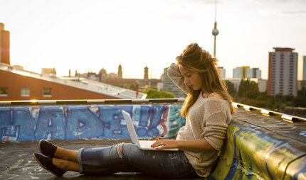 Junge Frau mit Laptop auf dem Schoß sitzt auf einem Dach
